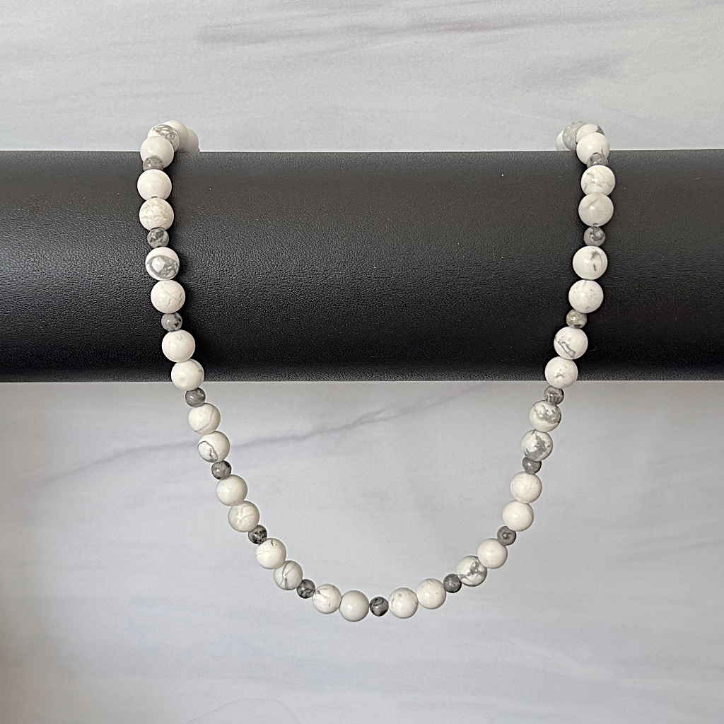 Mens Howlite and Map Beaded Necklace, draped over a black cylindrical display stand against a light gray marble-patterned background.
