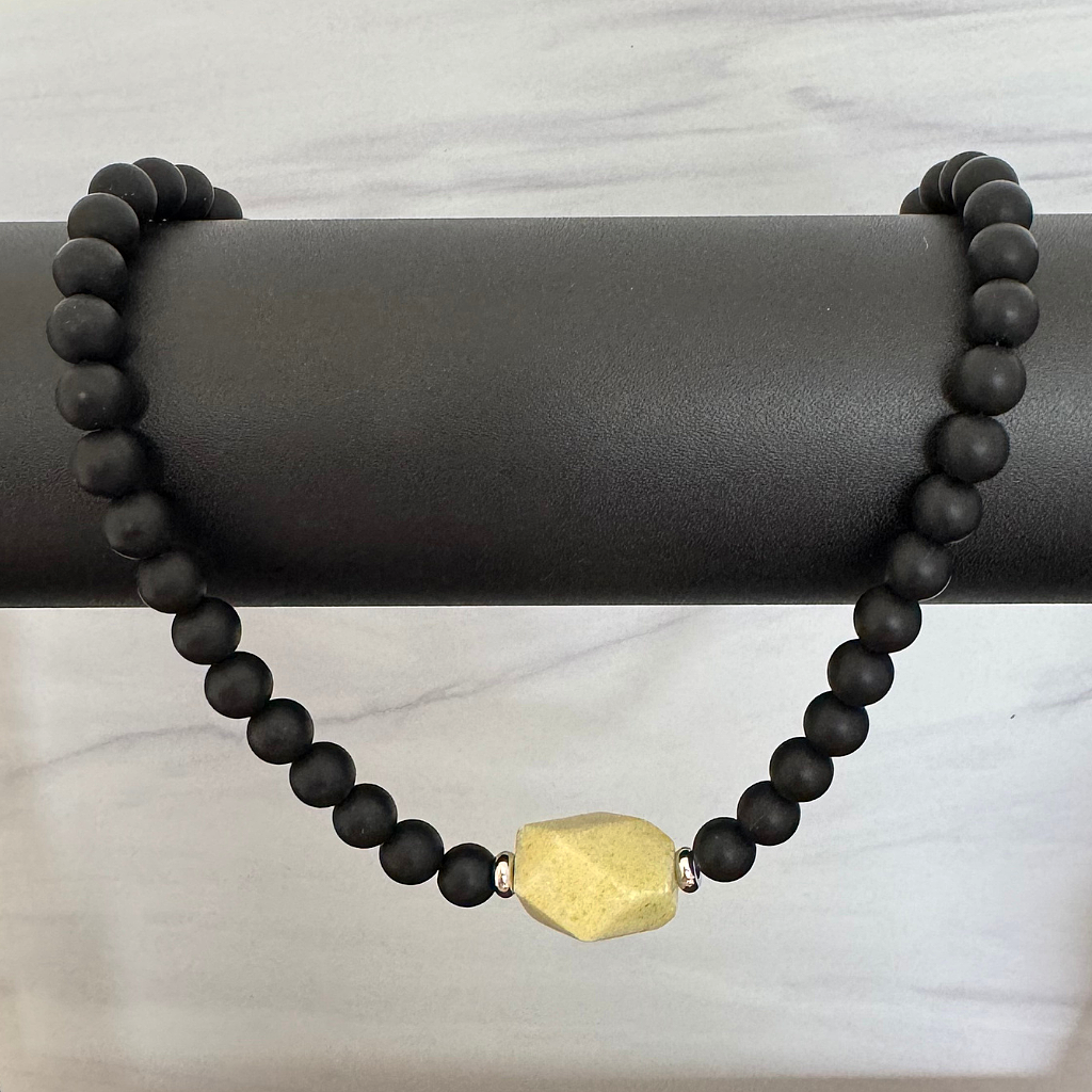 Black beaded necklace with a gold hexagonal bead on a dark cylindrical stand.