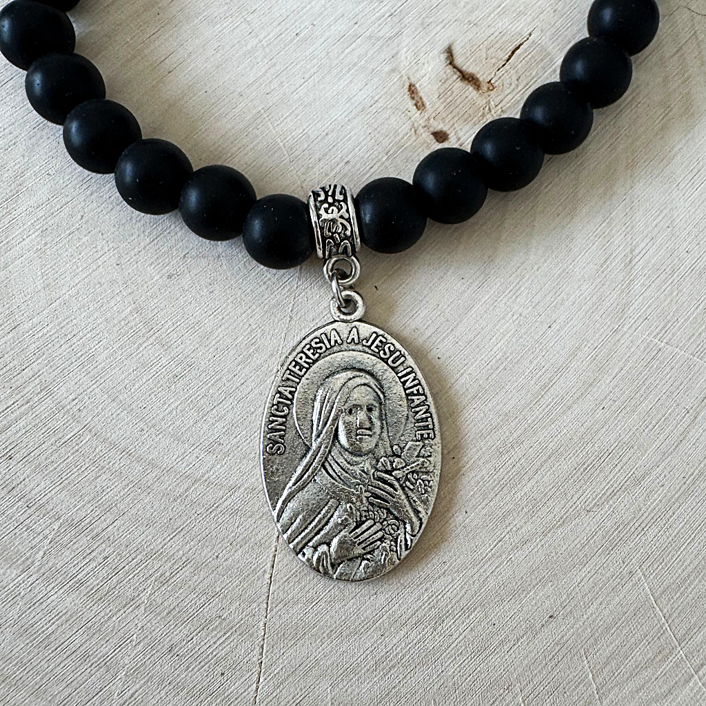 A beaded necklace with matte black onyx ound beads and a silver-tone oval Saint Teresa of Jesus pendant, displayed on a light beige textured fabric background.